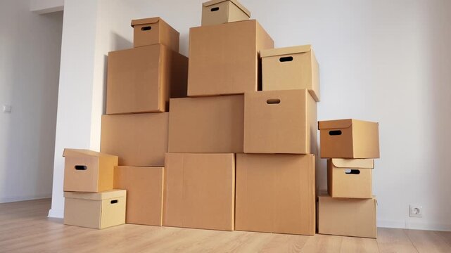Brown cardboard boxes stack in a bright room. Light wood floor and white walls provide clean background. Various sizes create balanced pyramid arrangement. Boxes suggest moving or storage setup
