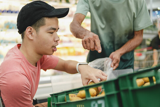 Caregiver teaching a young Asian man in a wheelchair how to select fresh potatoes in a supermarket, life skills learning, assistance, and inclusive development concept.