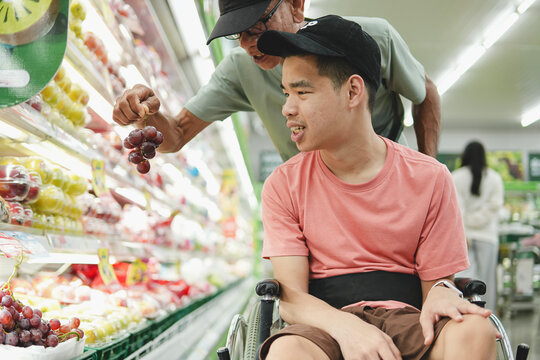 Young Asian man in wheelchair looking at fresh red grapes held by senior father in supermarket. Disabled shopper enjoying grocery shopping, family support, accessibility and inclusion concept.