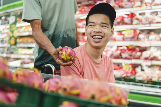 Happy young Asian man in wheelchair choosing fresh dragon fruit with father in supermarket. Disabled male customer enjoying grocery shopping, family support, accessibility and inclusion concept.