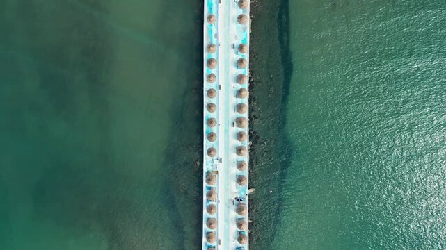Albania, Durres 07 09 2025: Top-down vertical drone video of a minimalist pathway over the sea connecting to Hawaii Beach Club, lined on both sides with straw umbrellas.