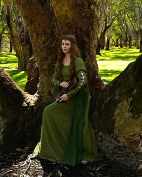 full length portrait of beautiful female model, sitting figure playing musical harp instrument, wearing green medieval historical gown costume with cloak. Magical forest background with trees.