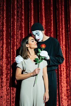 Mime artist with white face paint and black costume offers a red rose to a woman on theater stage. Woman accepts flower, they look at each other tenderly. Red velvet curtains backdrop, romantic scene.