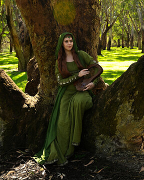 full length portrait of beautiful female model, sitting figure playing musical harp instrument, wearing green medieval historical gown costume with cloak. Magical forest background with trees.