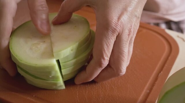 person cutting stack of zucchini slices in half with sharp knife. hands preparing zucchini halves for gratin with cheese at home. concept of zucchini cooking and homemade vegetable dish in kitchen