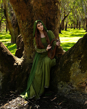 full length portrait of beautiful female model, sitting figure playing musical harp instrument, wearing green medieval historical gown costume with cloak. Magical forest background with trees.
