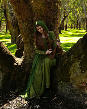 full length portrait of beautiful female model, sitting figure playing musical harp instrument, wearing green medieval historical gown costume with cloak. Magical forest background with trees.