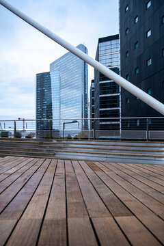 An empty office terrace extends across a deck beside corporate buildings, while a railing follows the urban walkway against surrounding architecture