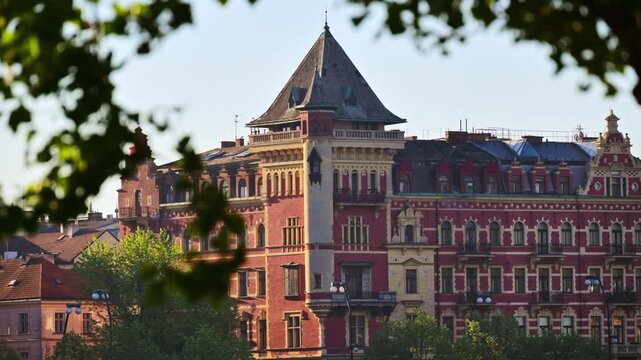 A red brick corner building with a pale stone tower and steep slate roof seen through green leaves, Prague, Czech Republic
