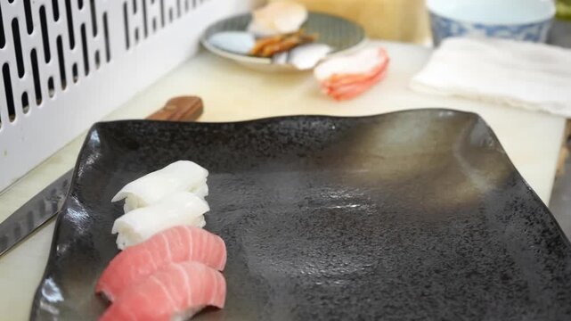 Professional chef arranges multiple types of fresh seafood nigiri sushi on a dark plate, adding red roe and side garnishes in a clean kitchen