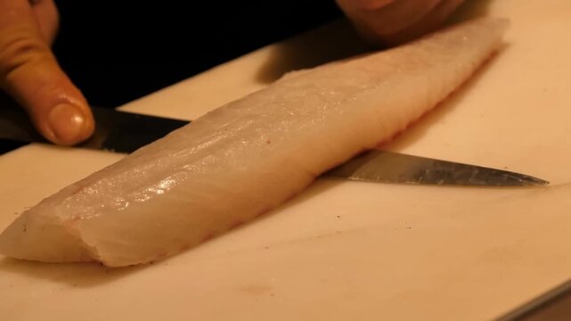 A professional chef utilizes a sharp knife to carefully fillet fresh raw fish on a white cutting board inside a commercial restaurant kitchen environment for meal preparation