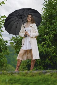 Woman stands in rain holding black umbrella. She wears white dress and light coat. Background shows green trees under rainy sky