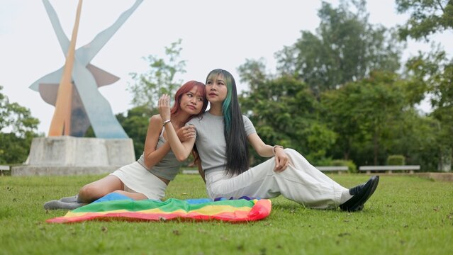 Smiling Asian women sitting together on lush grass field after outdoor picnic, embracing fondly with cheerful satisfaction, youthful freedom, and sweet togetherness in nature.