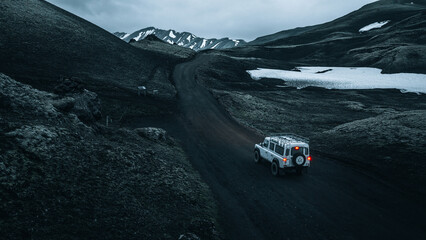 Obraz premium An off-road vehicle travels through the remote volcanic highlands near Mælifell, surrounded by black lava fields, lingering snow and dramatic mountain scenery in Iceland's untouched wilderness.