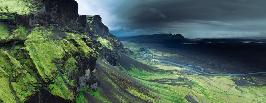 Towering moss-covered volcanic cliffs rise above a vast valley with braided rivers as dramatic storm clouds sweep across the rugged Icelandic landscape in breathtaking light.