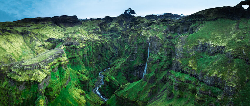 Panoramic aerial view of the lush M&uacute;laglj&uacute;fur Canyon with its hidden waterfall, towering moss-covered cliffs and winding river, one of Iceland's most spectacular natural landscapes.