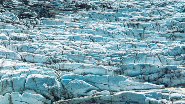Aerial close-up of ancient blue glacier ice revealing deep crevasses, dark volcanic ash layers and intricate frozen patterns at Skaftafellsj&ouml;kull in southeast Iceland.