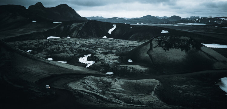 An off-road vehicle drives through the volcanic landscape of Landmannalaugar, surrounded by ancient lava fields, volcanic craters and scattered snow beneath dramatic clouds in Icelandic Highlands.