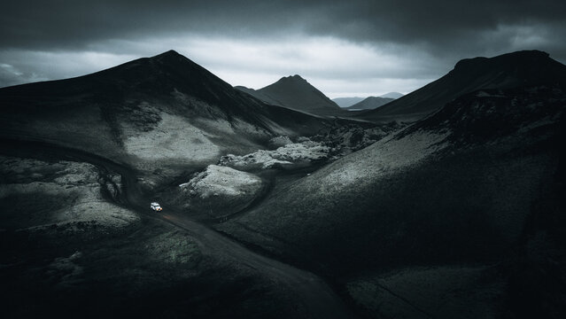 An off-road vehicle drives through the volcanic landscape of Landmannalaugar, surrounded by ancient lava fields, volcanic craters and scattered snow beneath dramatic clouds in Icelandic Highlands.