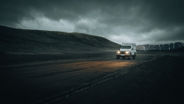 An off-road vehicle drives across the remote volcanic Highlands of Iceland beneath dark storm clouds, with headlights illuminating the rugged track through an untouched wilderness.
