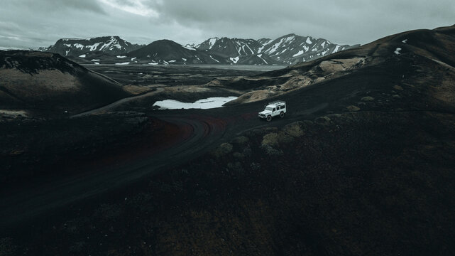 An off-road vehicle travels through the remote Icelandic Highlands near M&aelig;lifell, passing volcanic mountains, lingering snowfields and rugged lava terrain beneath dramatic overcast skies.