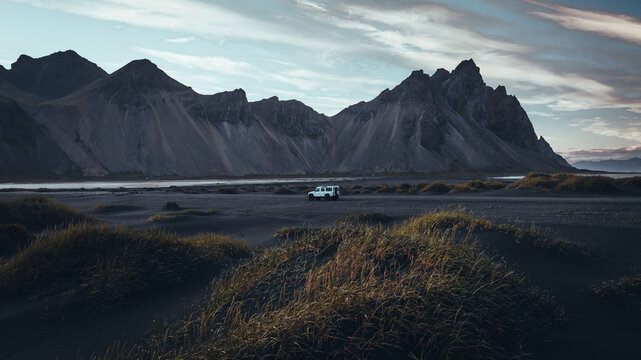 An off-road vehicle stands on the black sand plains of Stokksnes beneath the dramatic peaks of Vestrahorn, surrounded by windswept grass, rugged coastline and evening light in Iceland.