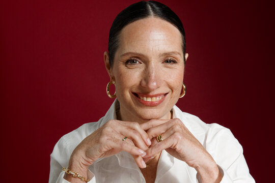 Woman Smiles With Hands Clasped in Front of Red Background