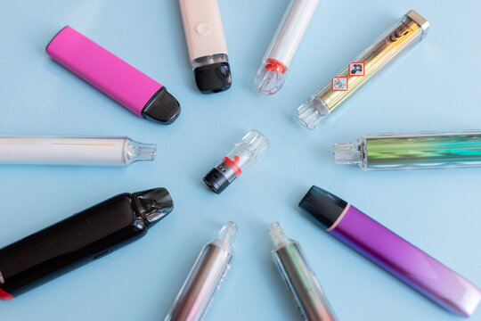 Modern electronic vaping devices of various shapes and colors surrounding a single toxic product. A striking display of a health threat.