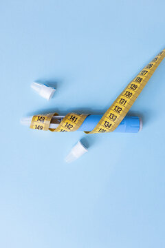 Vertical shot of blue weight loss injection pen wrapped in yellow measuring tape on blue background, illustrating extreme dieting risks, medication misuse, side effects, and health hazards.