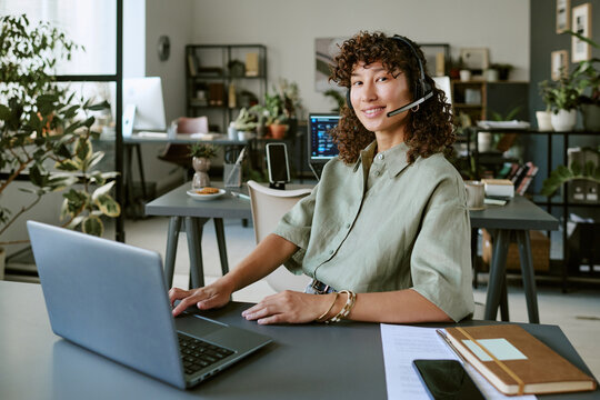 Portrait of smiling young adult Biracial woman wearing headset working on laptop in modern office. Image represents customer support and is suitable for corporate communication