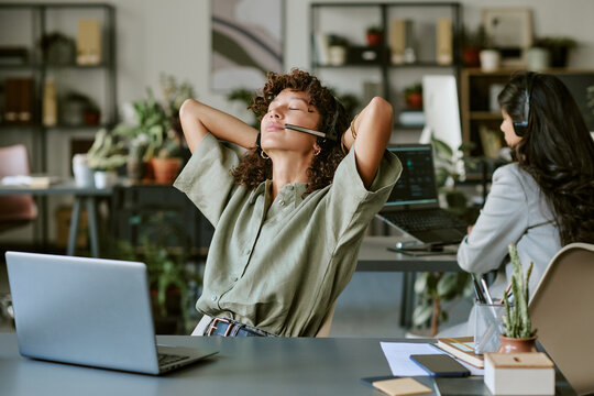 Diverse young adult female call center operators working in office, one stretching with closed eyes. Image represents mental wellbeing and stress relief in corporate communication