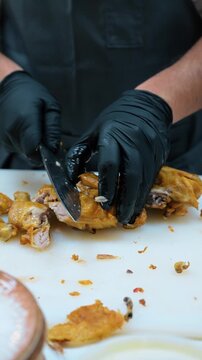 Cook cutting browned chicken with cleaver. Black gloves hold a poultry portion on a white board scattered with skin and small scraps.