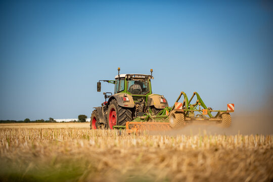 A tractor pulling a cultivator kicks up dust while working a wheat stubble field under a clear blue sky in the Eure region, Normandy, France, captured with a low angle and shallow depth of field.