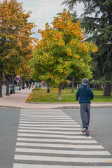 Fototapeta premium Person Riding Electric Scooter Across Urban Crosswalk in Autumn. Young man riding an electric scooter, pedestrian crossing, autumn city street. Urban micromobility, sustainable personal transportation