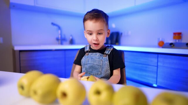 Young boy holding bitten yellow apple at counter in blue illuminated kitchen. Rows of fresh fruit rest on a white surface in front of a child wearing denim overalls.