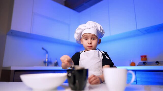 Young boy wearing toque and apron at kitchen table with white cookware. A child plays chef behind blurred dishes on a bright counter under blue lighting.