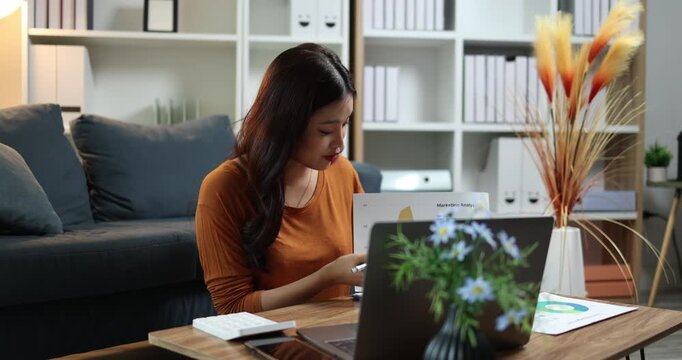 Young asian woman working remotely on laptop and phone in modern home living room, freelancer productivity