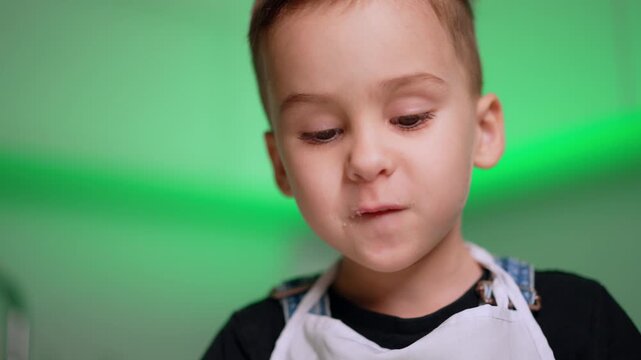 Young boy wearing white apron looking down in portrait. A child licks a crumb from his mouth against a bright green background.