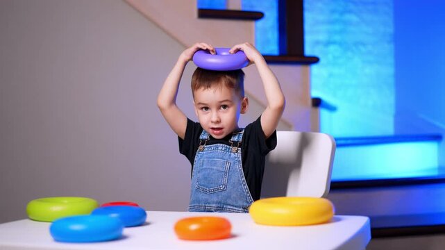 Young boy holding purple ring toy on head at table. A child sits with colorful stacking rings on a white desk indoors.