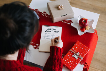 Fototapeta premium Woman writes in notebook with HOLIDAY BUDGET PLAN on table beside gifts and COZY CHRISTMAS sign. Saving money for christmas, financial goal setting holiday, christmas expense management