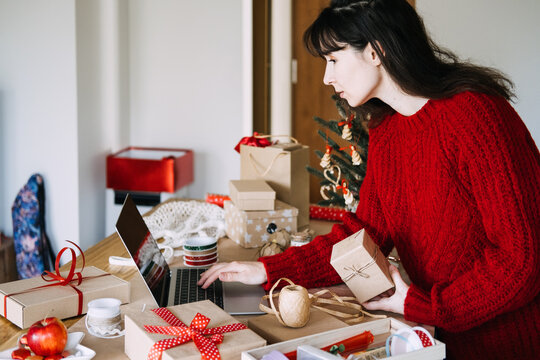 Woman holds kraft wrapped parcel while working on laptop among gifts and wrapping supplies. Christmas small business laptop, managing orders christmas season, small business holiday computer work