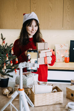 Smiling woman in Santa hat holds stack of wrapped parcels before phone on tripod in kitchen. Small business packing orders, handmade gift packaging video, etsy seller holiday content