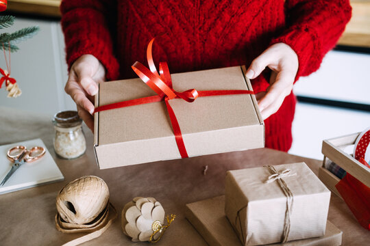 Woman hands hold kraft box tied with ribbon above table with wrapping supplies. Mindful gift giving concept, quality over quantity christmas, intentional consumerism holiday