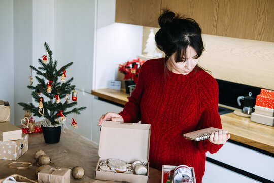 Woman holds notebook while checking open box with brush and jar in kitchen. Small business checking order list, verifying christmas order contents, matching gift to checklist