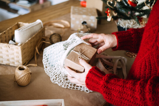 Hands place kraft wrapped parcel into mesh bag on table beside twine and small tree. Cozy christmas preparation home, festive wrapping session at home, christmas prep warm atmosphere
