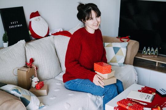 Smiling woman holds stack of wrapped boxes on sofa beside notebook with HOLIDAY BUDGET PLAN and COZY CHRISTMAS sign. Holiday budget planning notebook, personal finance christmas season, 