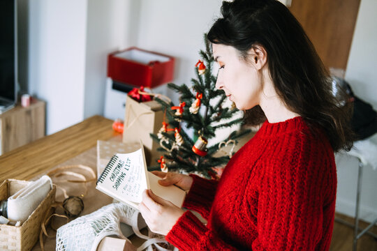 Woman reads notebook with CHRISTMAS SHOPPING LIST beside decorated tree and wrapping supplies. Checking christmas shopping list, reviewing gift list before shopping, christmas shopping list checklist