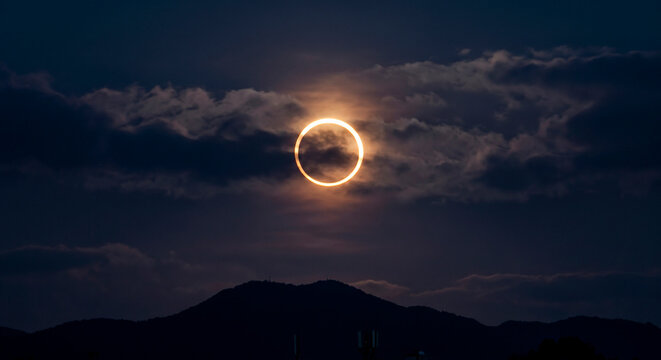 Espectacular eclipse solar anular con el deslumbrante "anillo de fuego" entre nubes oscuras, sobre la silueta de una monta&ntilde;a al anochecer.