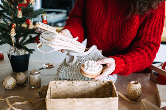 Woman holds mesh bag and rolled cotton towel above basket with gift supplies on table. Eco friendly christmas gift set, sustainable home care package christmas, zero waste christmas basket