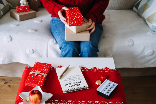 Woman holds stack of wrapped boxes on sofa above table with HOLIDAY BUDGET PLAN notebook and HOLLY JOLLY sign. Holiday budget planning notebook, personal finance christmas season, tracking christmas